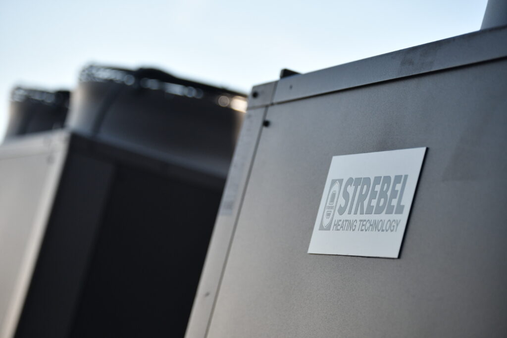 Close-up of a Strebel commercial air source heat pump casing showing the brand logo, with large extraction fans visible in the background.